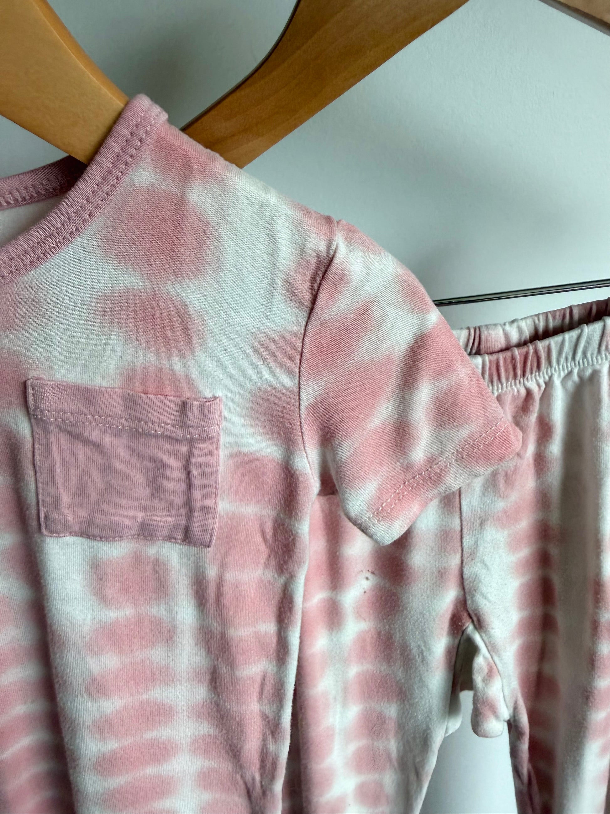 Pink and white tie-dye short-sleeve pajama top with a chest pocket, hanging on a wooden hanger next to matching tie-dye pants.